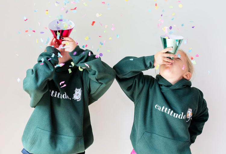 Two girls wearing green Cattitude hooded sweatshirt from The Joyful Rebel. The two girls are blowing confetti in to the air.