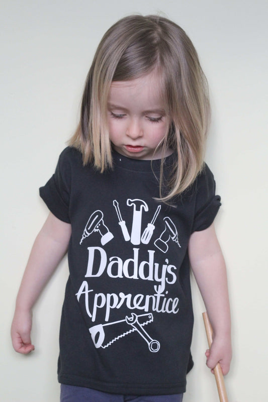 Blonde toddler girl wearing a black t-shirt with white logo 'Daddy's Apprentice'