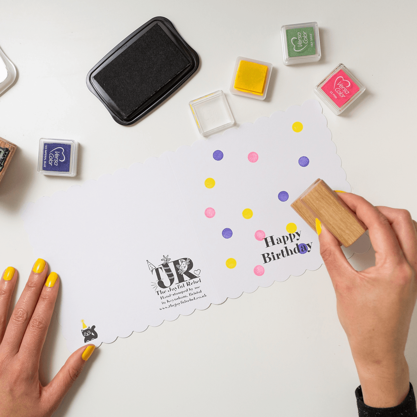 Person using a stamp on a card with colorful dots and 'Happy Birthday' text, surrounded by ink pads.