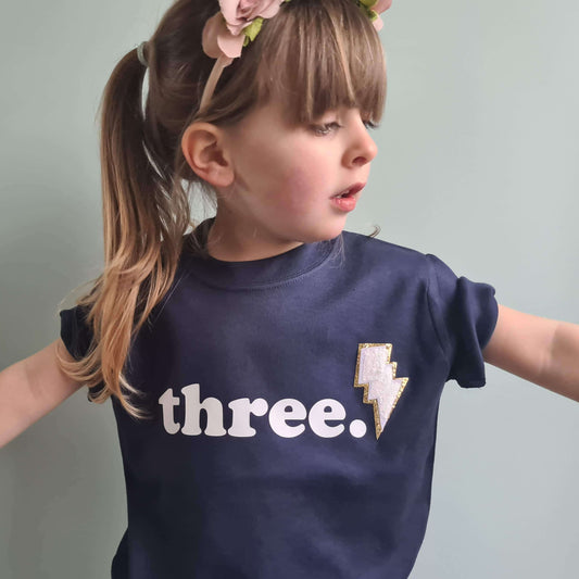 girl wearing a navy blue 3rd birthday tshirt. The t-shirt has white text 'three' and glitter lightning bolt