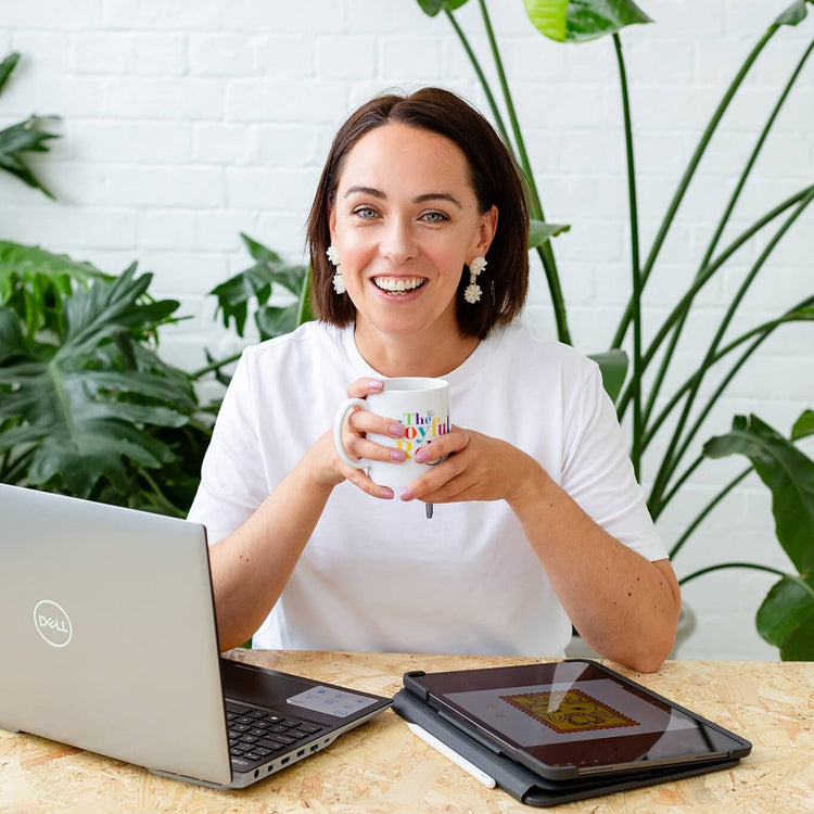 A woman smiling and holding a white ceramic mug with colorful text, seated at a desk with a laptop, tablet, and plant leaves in a bright indoor setting.