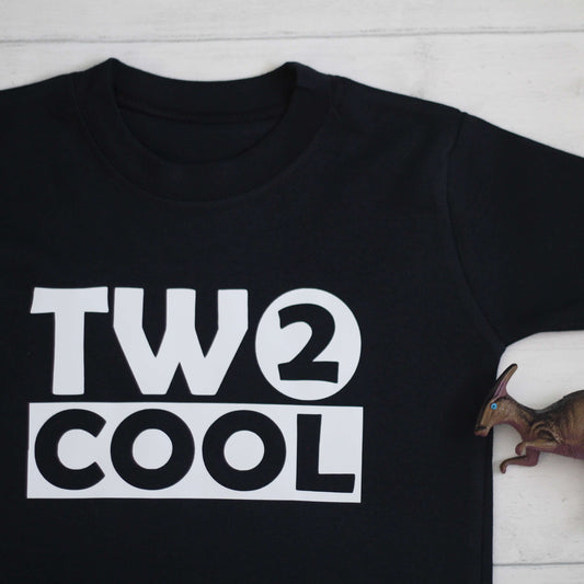 Black tshirt is laid on the floor. The top has a white logo with the text 'TWO COOL'