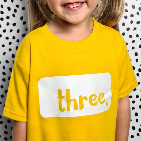 Toddler wearing a bright yellow birthday t-shirt with white logo 3 made by The Joyful Rebel