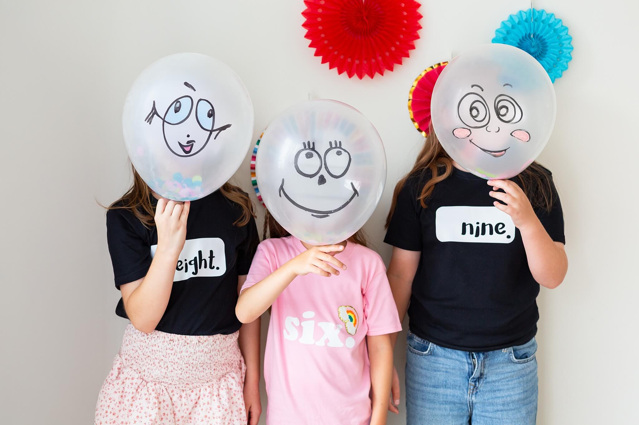 Three children holding balloons with faces drawn on, wearing t-shirts with numbers.