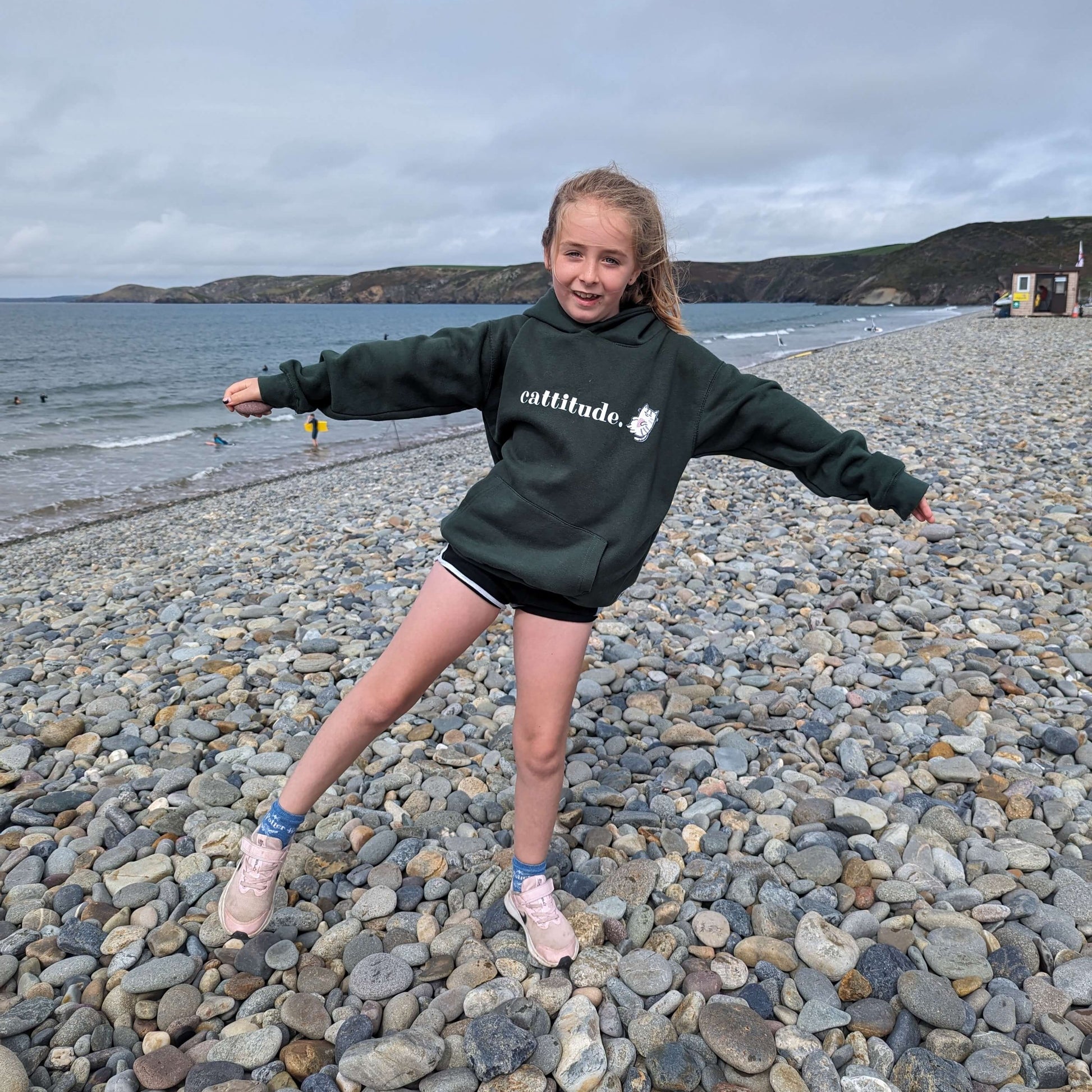 Girl wearing the Joyful Rebel green sweatshirt on a stoney beach.