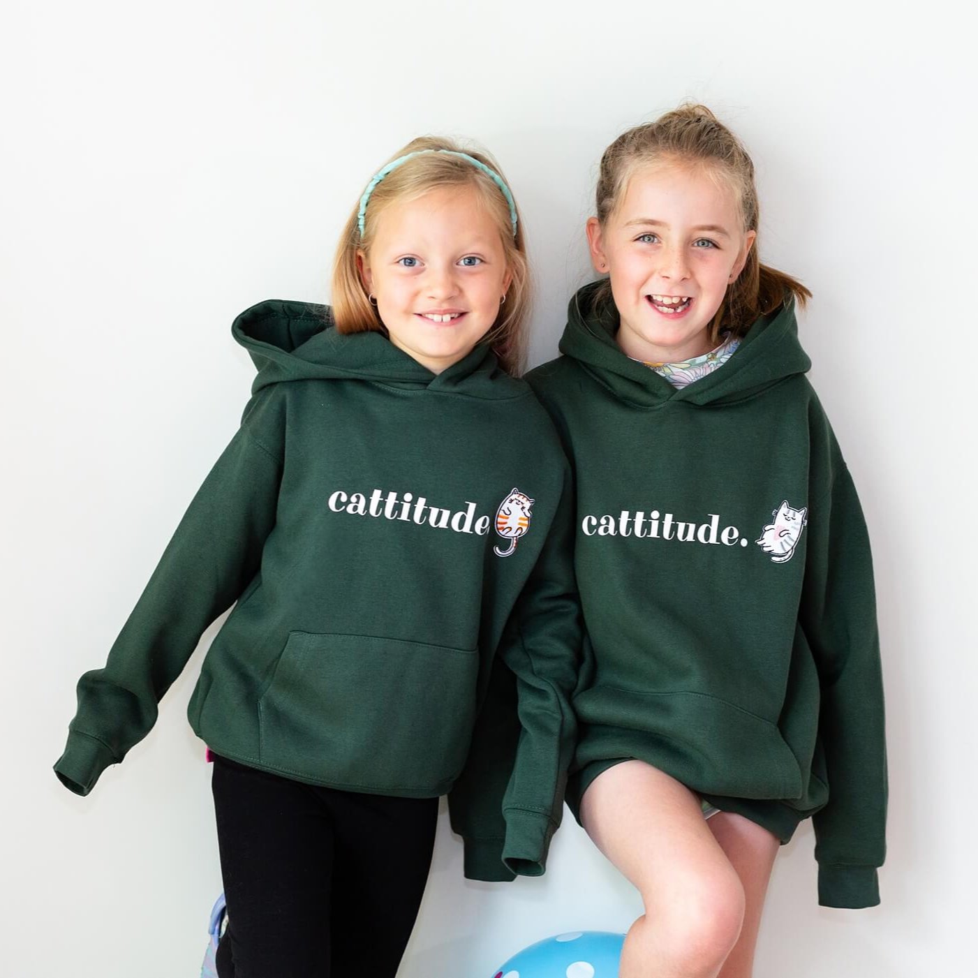 Two smiling girls wearing a green cat kids hoodie. Hoodie is made by The Joyful Rebel