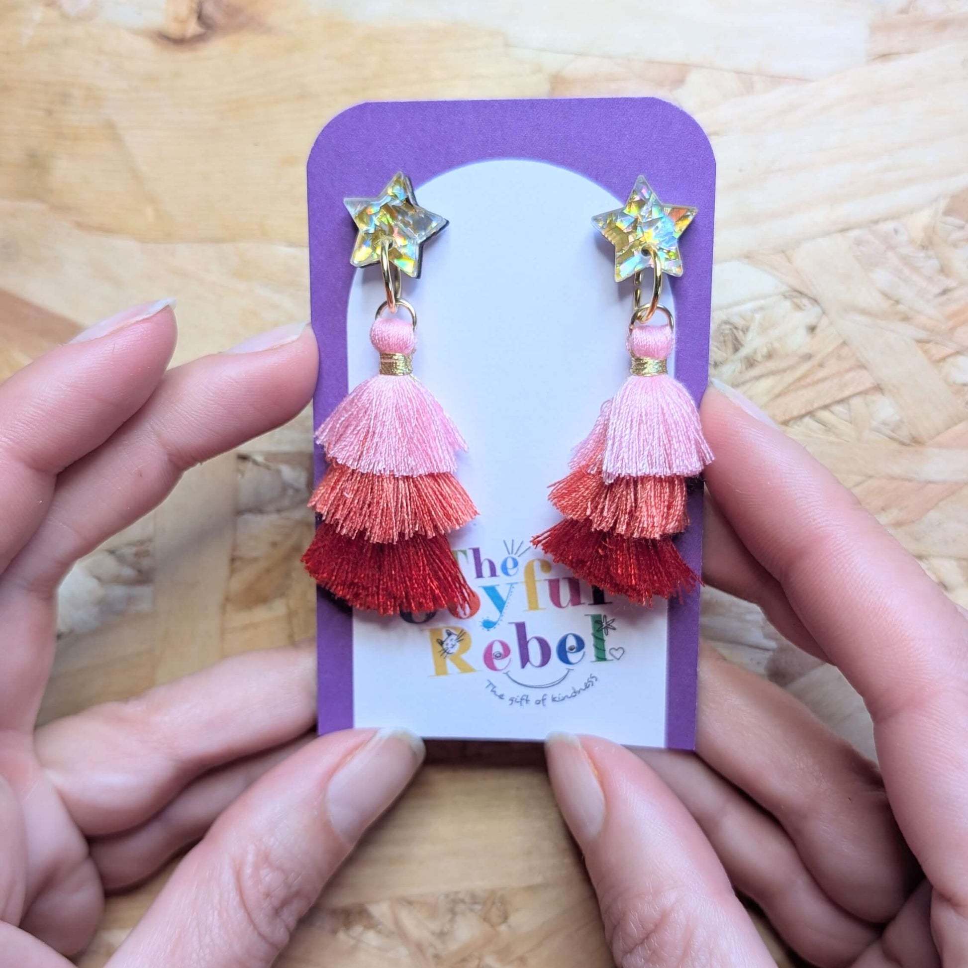 Colorful tassel earrings on a 'The Joyful Rebel' branded card held by hands on a wooden surface.