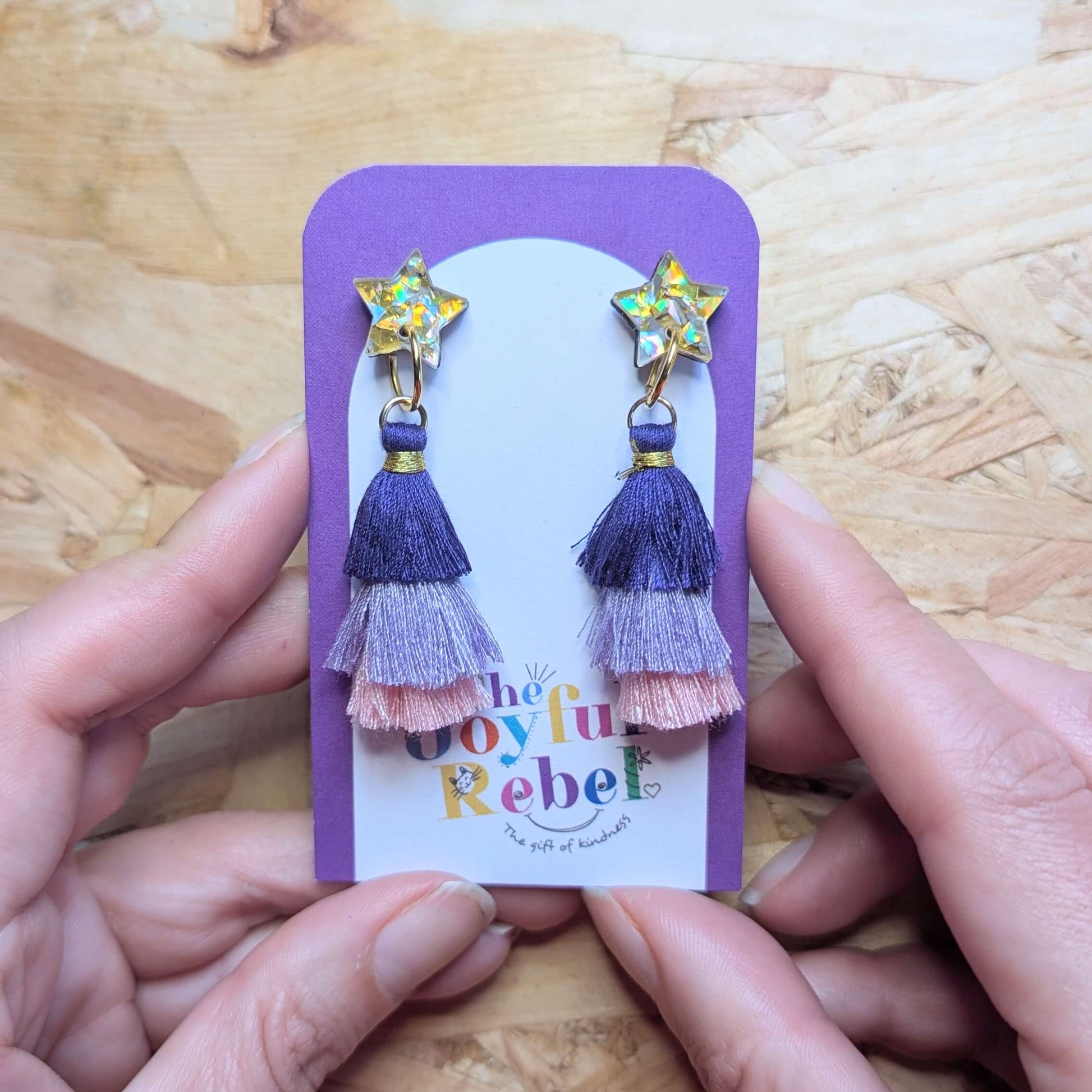 Purple and pink tassel earrings on a 'The Joyful Rebel' card held by hands on a wooden surface.