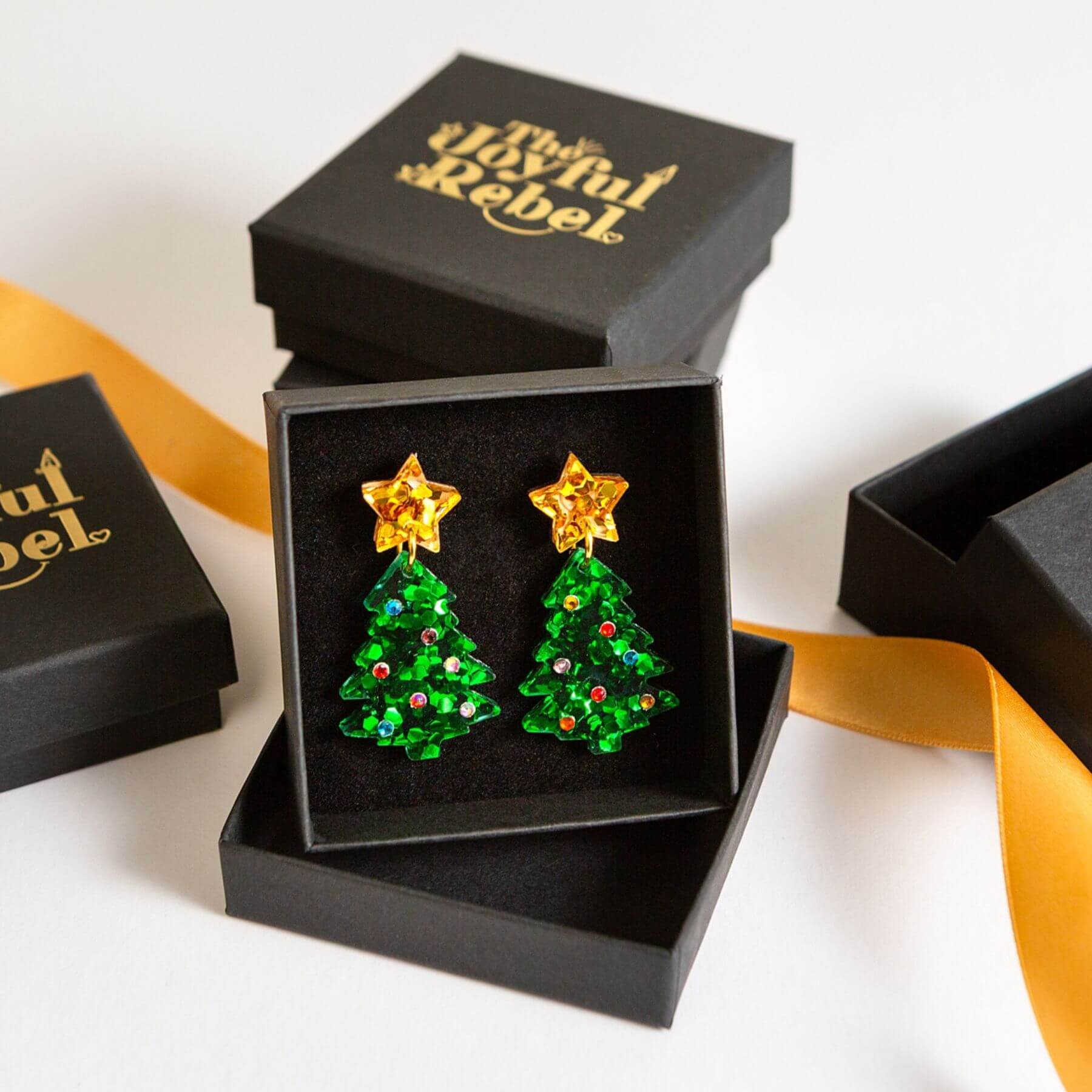 A pair of Christmas tree stud earrings with gold star tops and green sequined bodies, displayed in an open black gift box. 