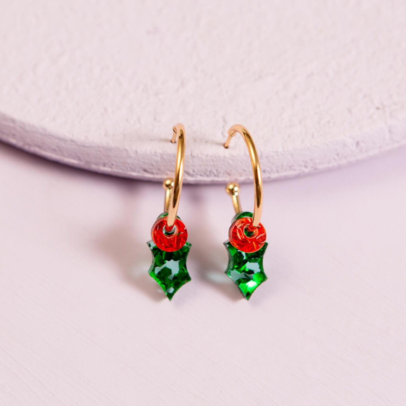 Gold earrings with red and green holly design on a light purple background