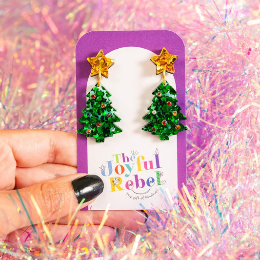 Green Christmas tree earrings on a purple card with 'The Joyful Rebel' branding against a glittery pink background.