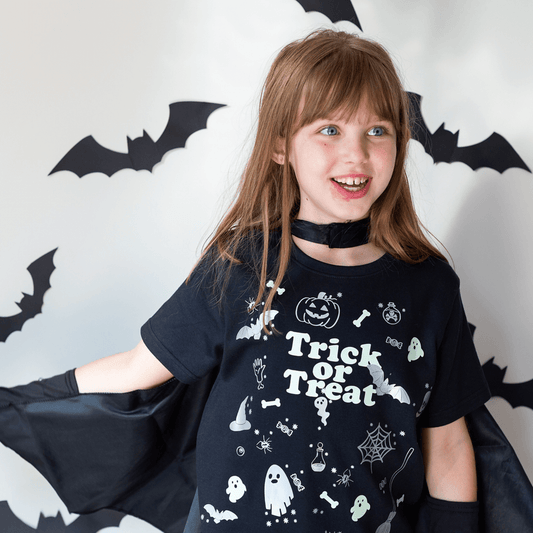 Child wearing a black 'Trick or Treat' shirt with Halloween designs against a decorative background with bats.
