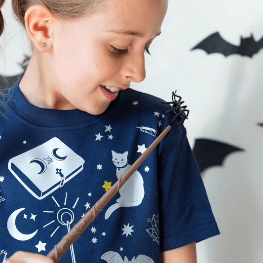 Child wearing a blue t-shirt with white magic graphics, holding a wand, with a white wall and black bat decorations in the background.