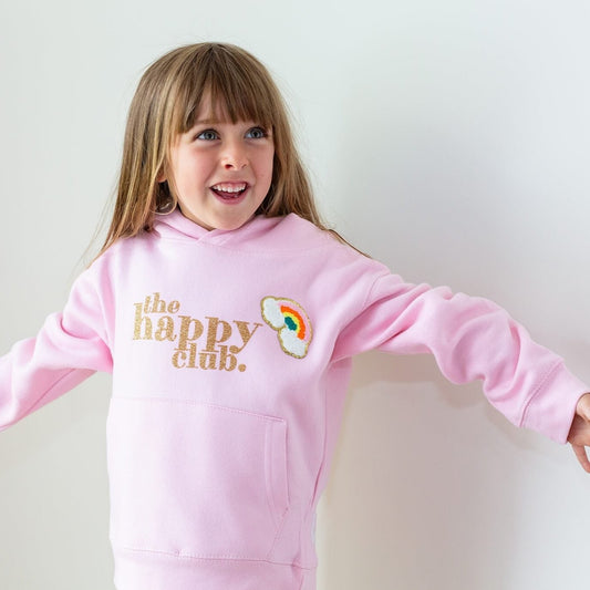 shows a young smiling girl wearing a pale pink hoodie. The glittery gold text reads 'the happy club' with embroidered rainbow