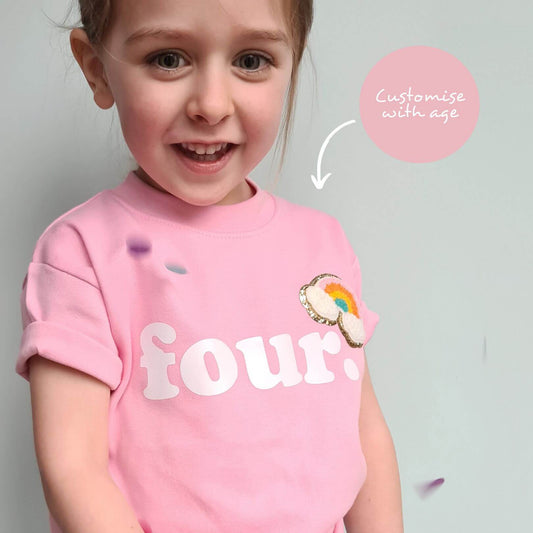 small child wearing a pastel pink tshirt. Pink tshirt has the text 'four' with an rainbow embroidered, gold edgine.