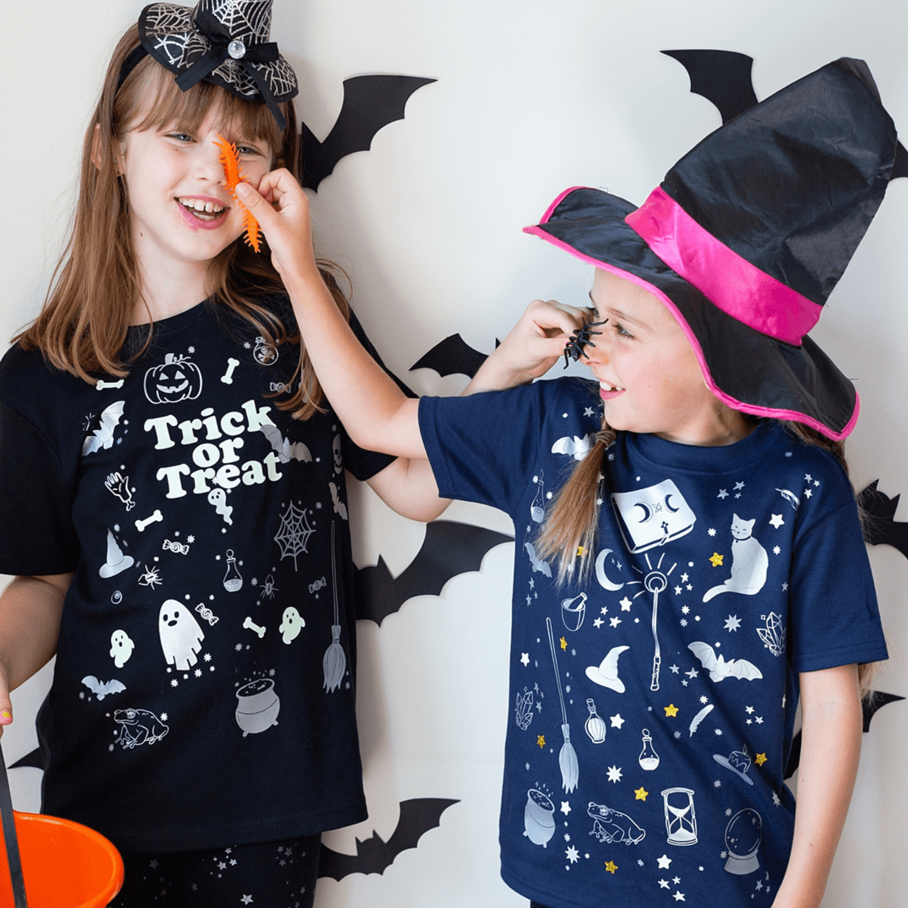Two children wearing Halloween-themed and wizard shirts with decorative bats in the background.