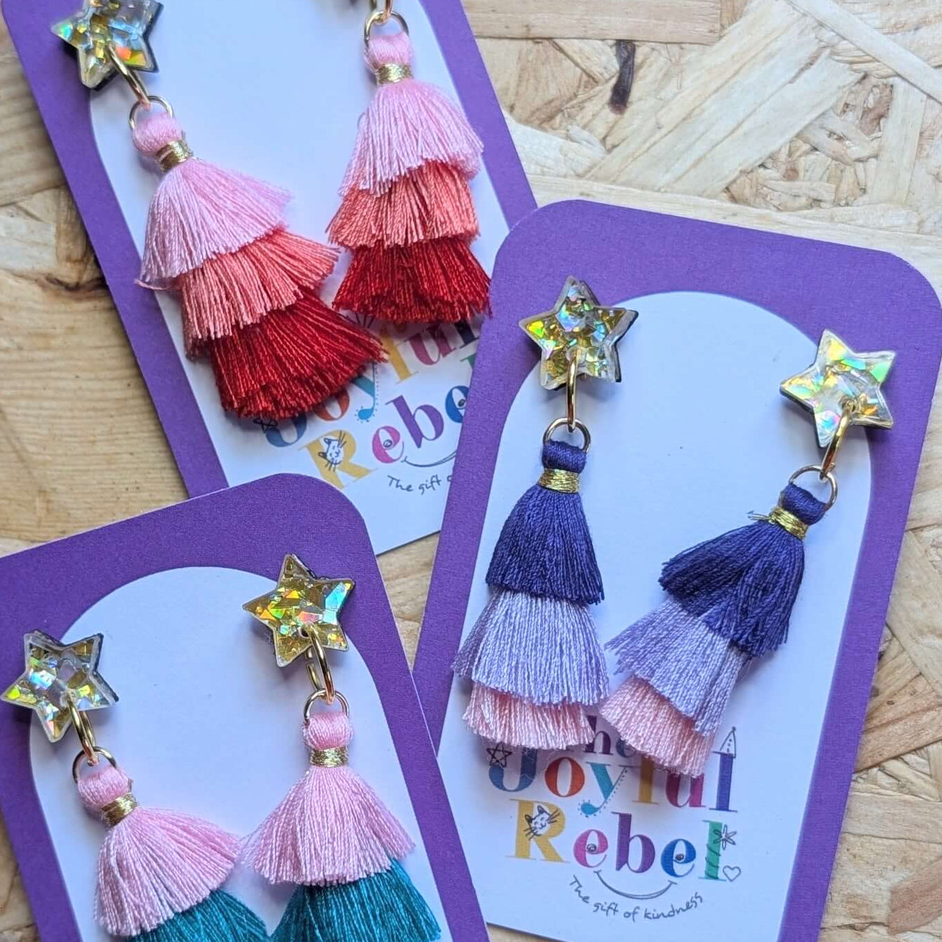 Colorful tassel earrings on purple cards with 'The Joyful Rebel' branding on a wooden surface.