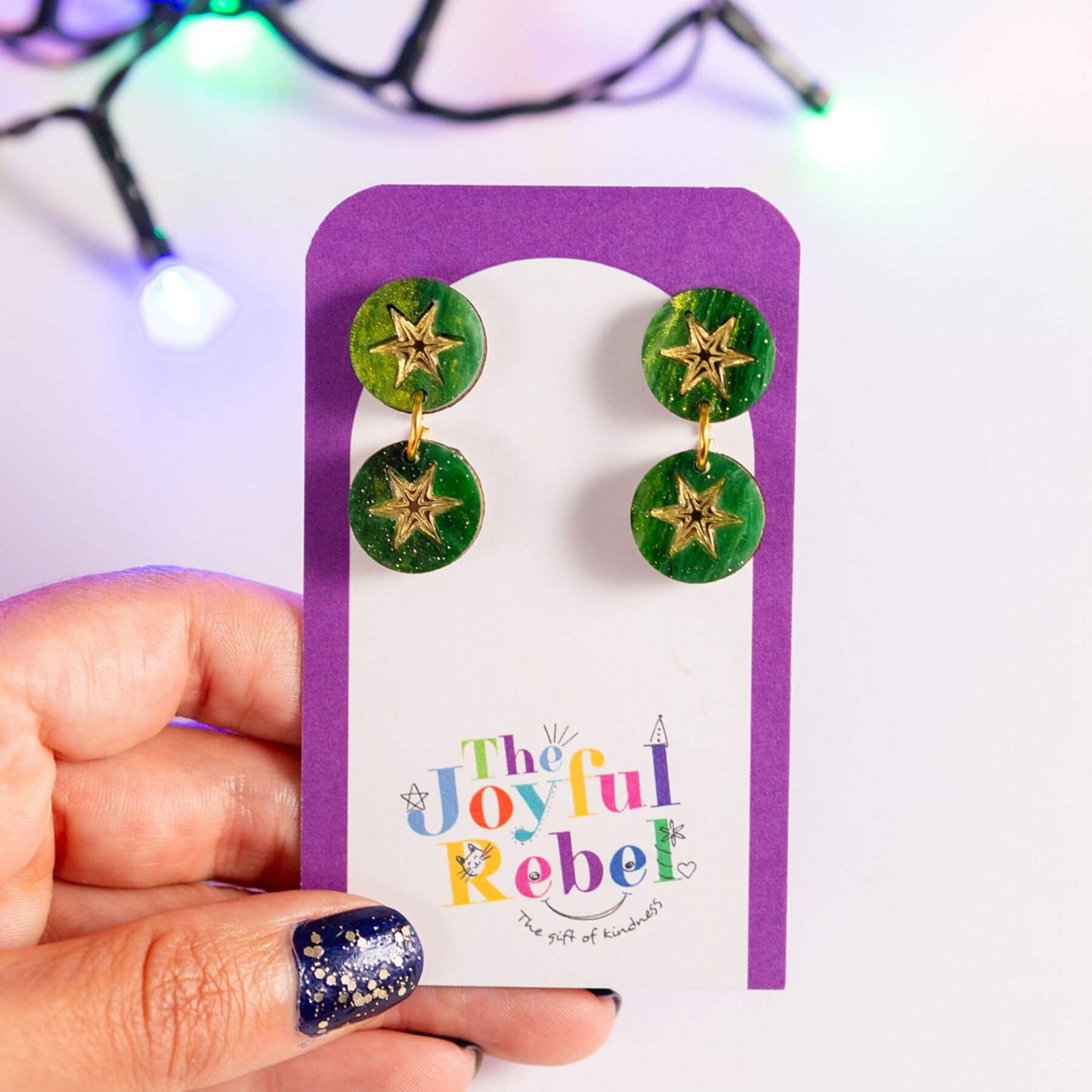 A hand holds a purple display card featuring a pair of green round drop earrings with gold star charms. The card is branded 'The Joyful Rebel' and is photographed against a bright white background with blurred colorful string lights.