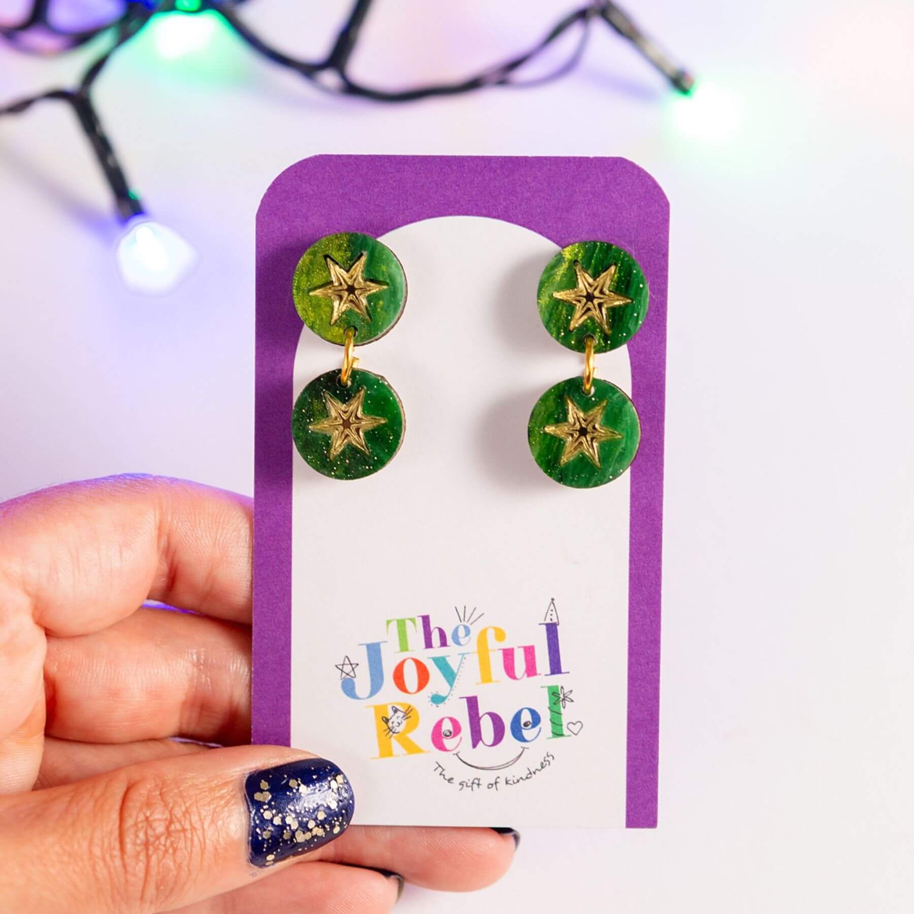 A hand holds a purple display card featuring a pair of green round drop earrings with gold star charms. The card is branded 'The Joyful Rebel' and is photographed against a bright white background with blurred colorful string lights.