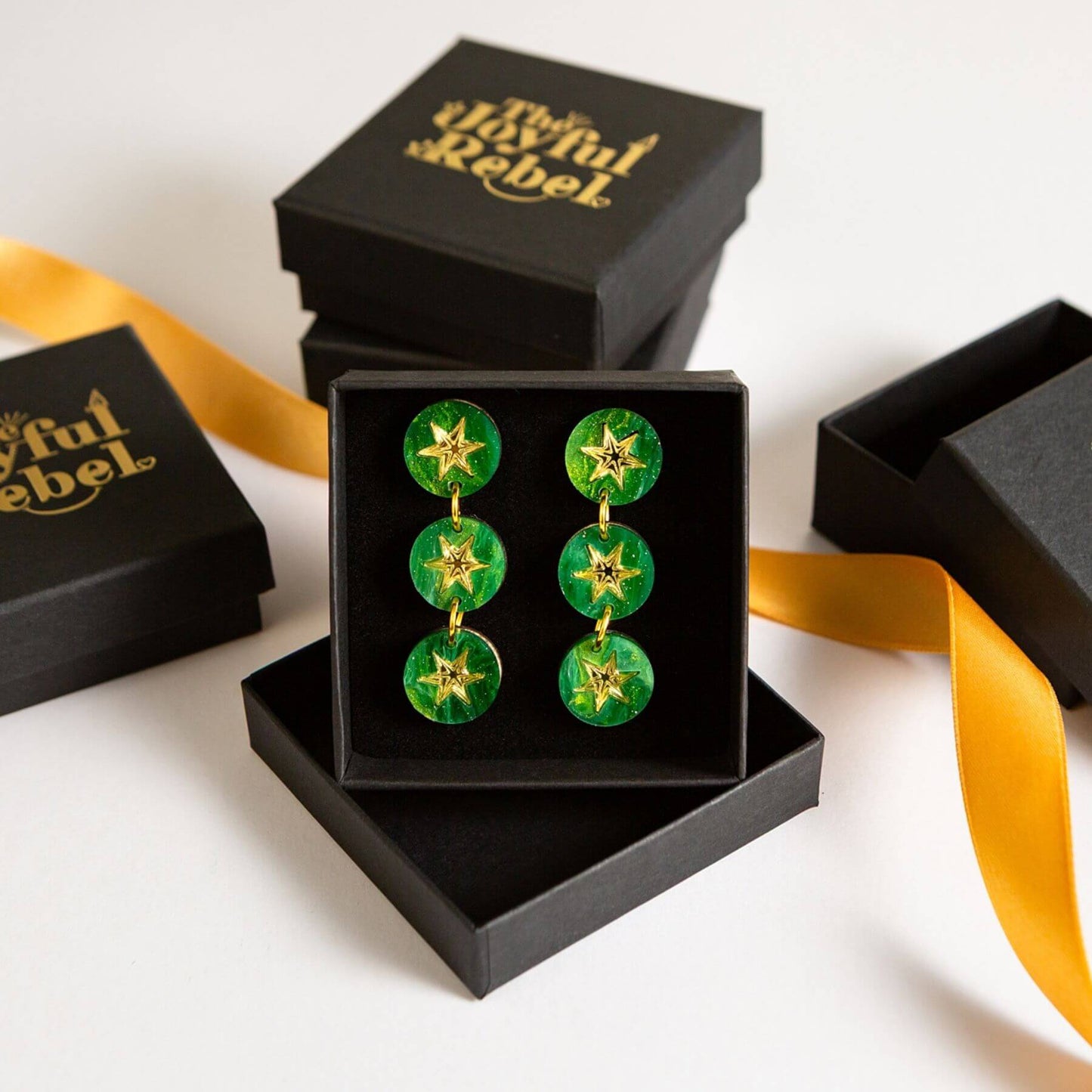 A pair of green and gold drop earrings displayed in an open black gift box, with additional black branded boxes and a yellow ribbon on a white background.