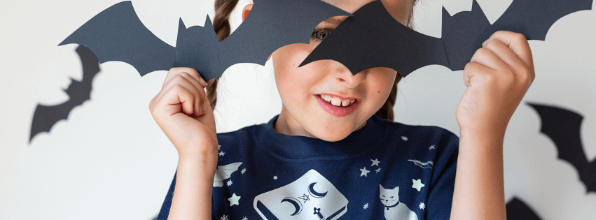 Child wearing a dark blue shirt with a wizard illustrations, holding black bat wings above their head against a light gray background.