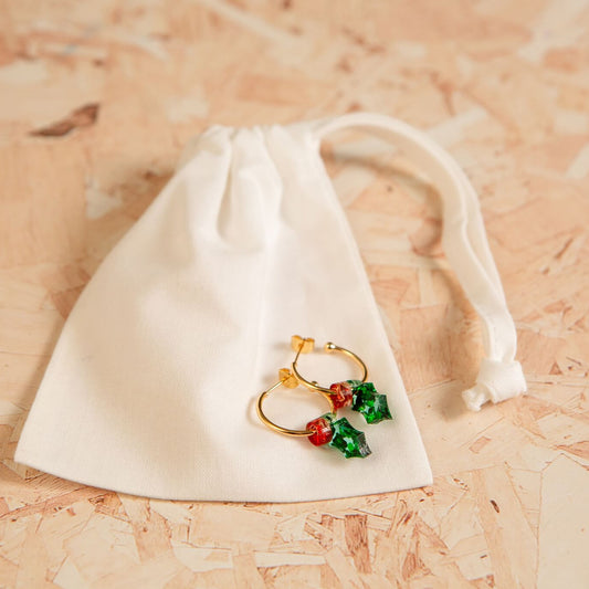 Gold and red holly earrings on a white drawstring bag against a wooden background