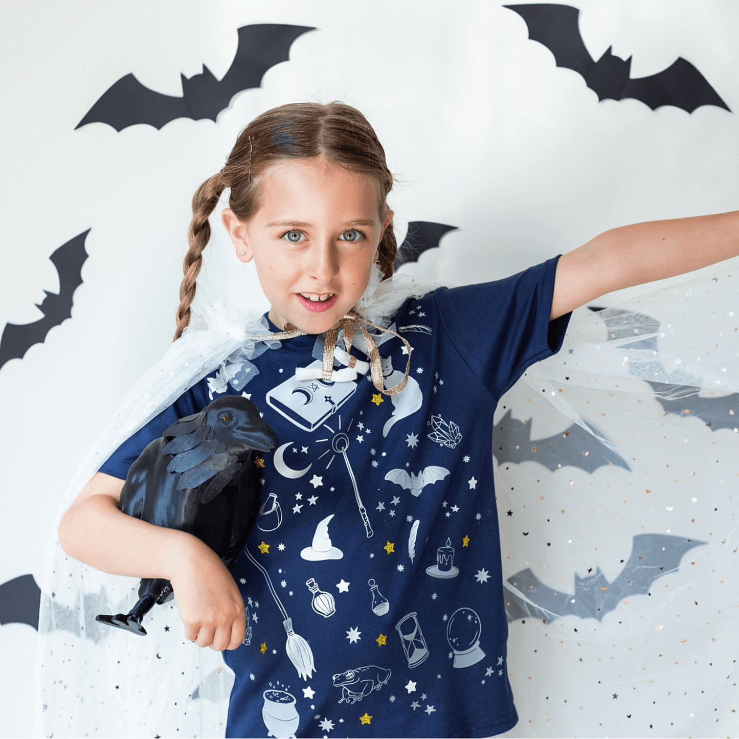 Child wearing a blue wizard-themed shirt with bats and a raven, standing against a white background with bat decorations.