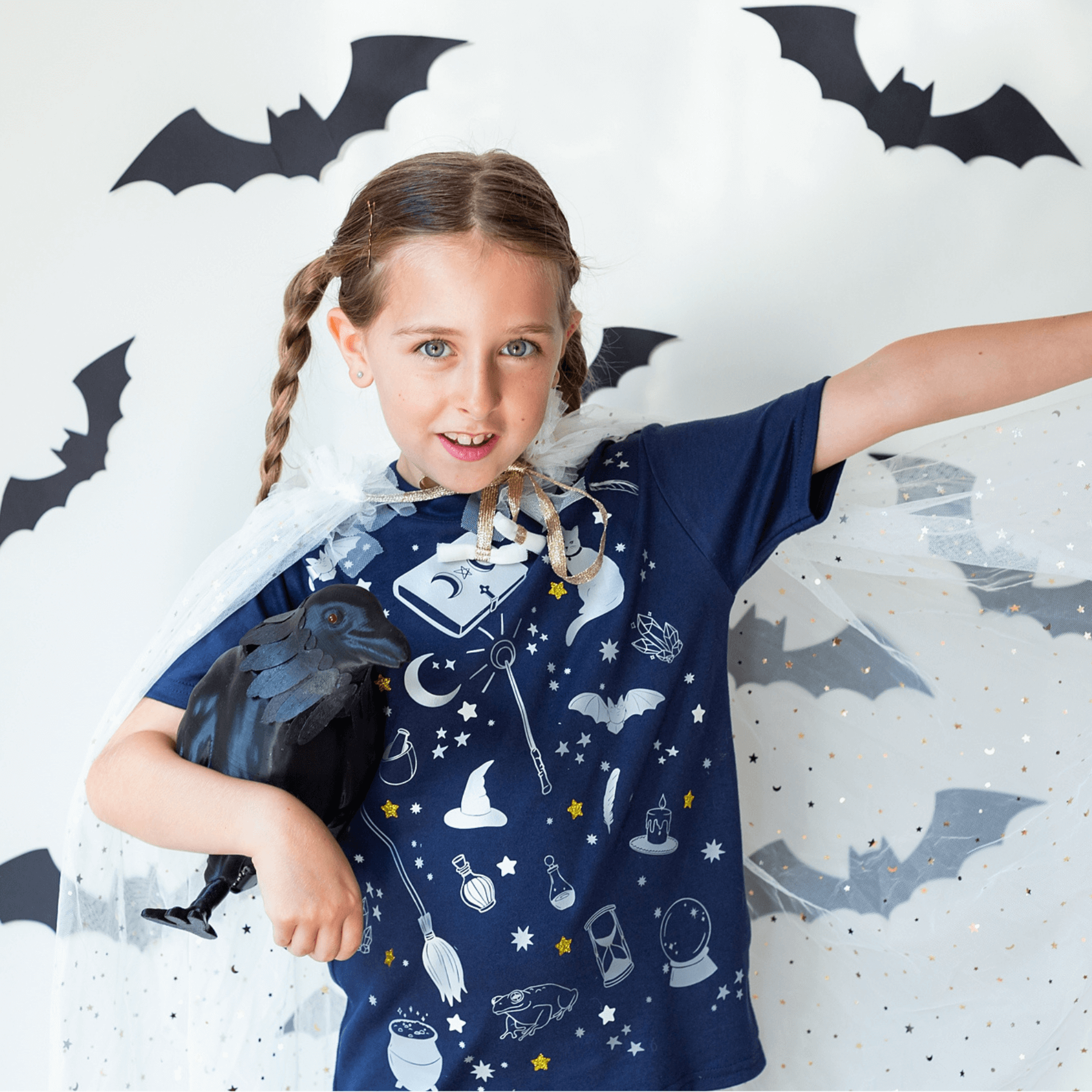 Child wearing a blue wizard-themed shirt with bats and a raven, standing against a white background with bat decorations.