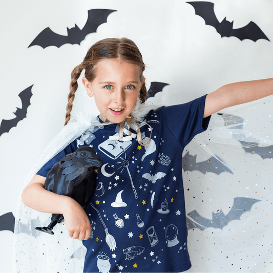 Child wearing a blue wizard-themed shirt with bats and a raven, standing against a white background with bat decorations.